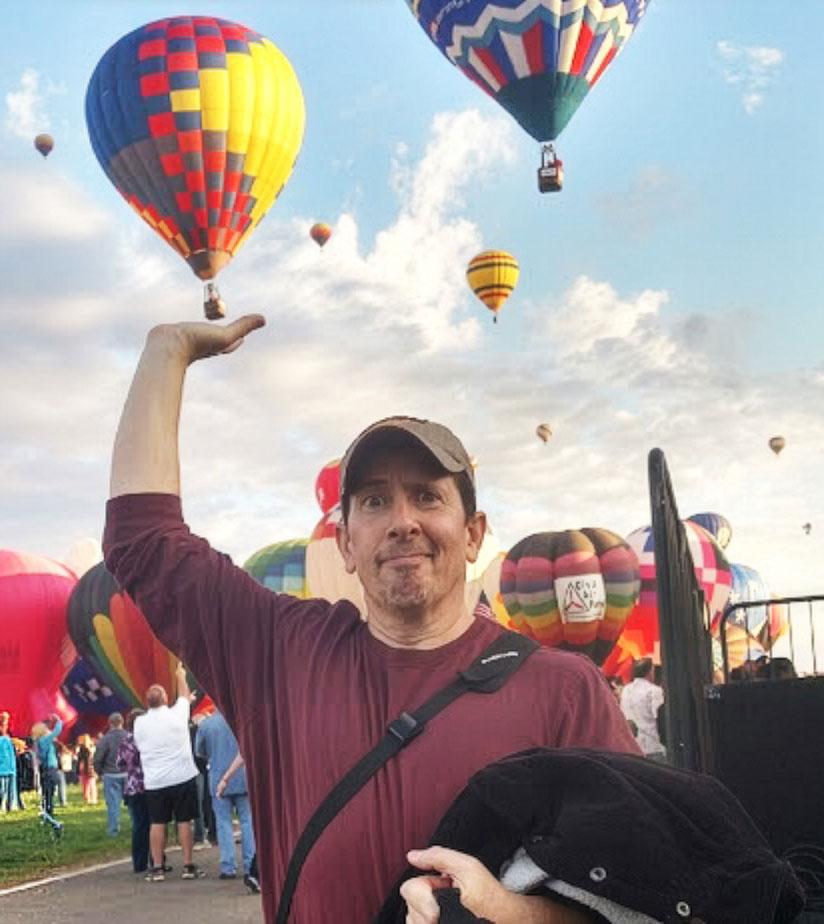 Weird man pretending to hold up a hot air balloon. Like we haven't seen that before. Whatever. Man with a baseball cap holding his arm up as if supporting a hot air balloon in Albuquerque NM.